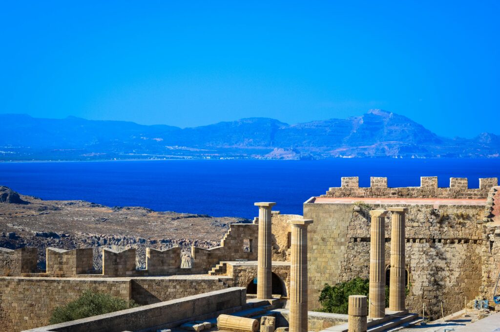 Scenic view of ancient Greek ruins with stone columns and walls against a vibrant blue Aegean Sea backdrop.