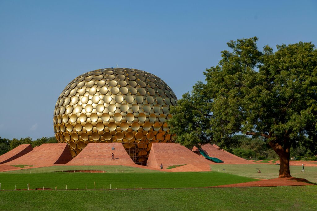 The iconic Matrimandir in Auroville, Puducherry, surrounded by green lawns and a vibrant tree under a clear blue sky.